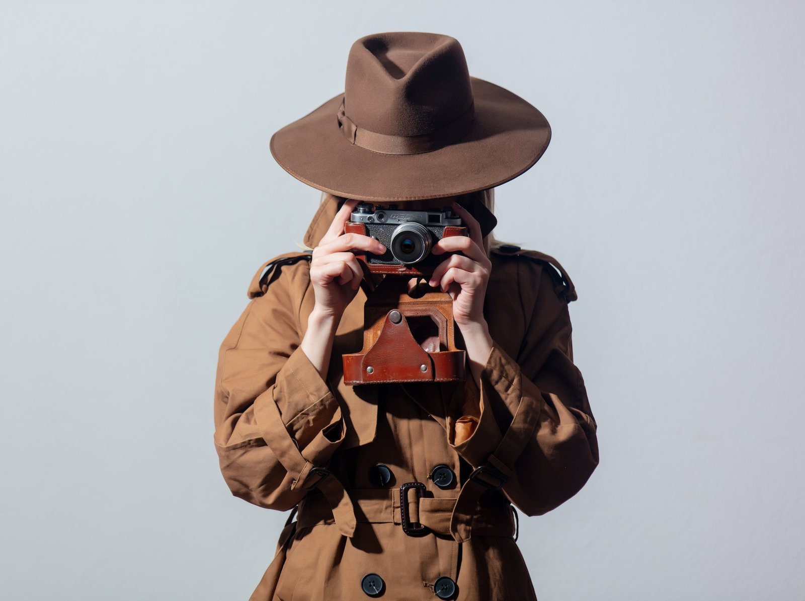 Woman in hat and cloak as a spy with camera on gray background.